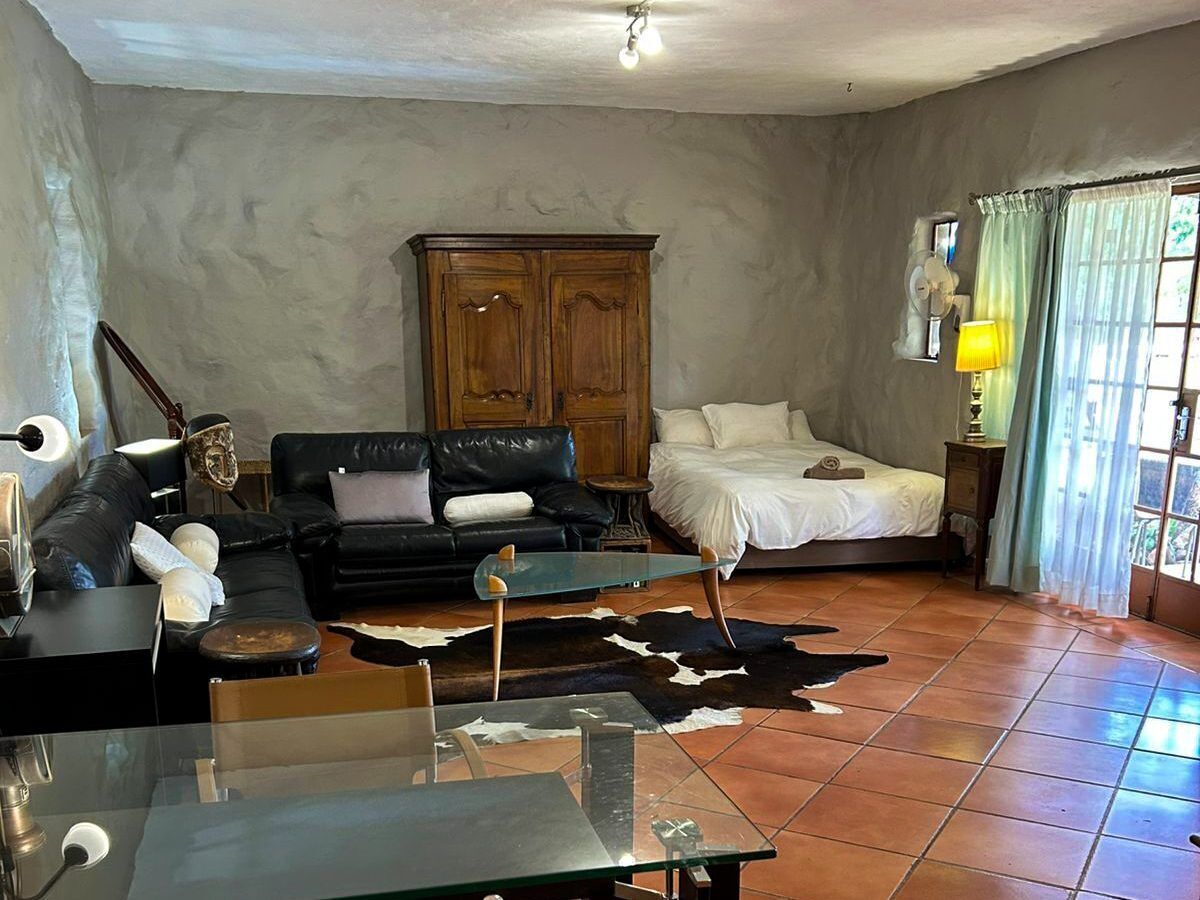 A living room with a black leather sofa, a glass coffee table, a wooden cabinet, a bed with white bedding, and a cowhide rug.