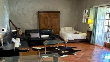 A living room with a black leather sofa, a glass coffee table, a wooden cabinet, a bed with white bedding, and a cowhide rug.
