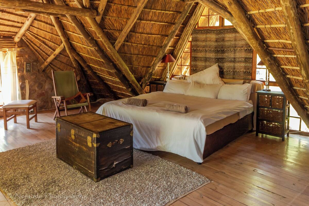 A rustic bedroom with a high wooden ceiling, a large bed, a wooden chest, and a stone wall.