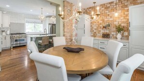A modern kitchen with white cabinets, a brick accent wall, a chandelier, and a round wooden dining table with white chairs.