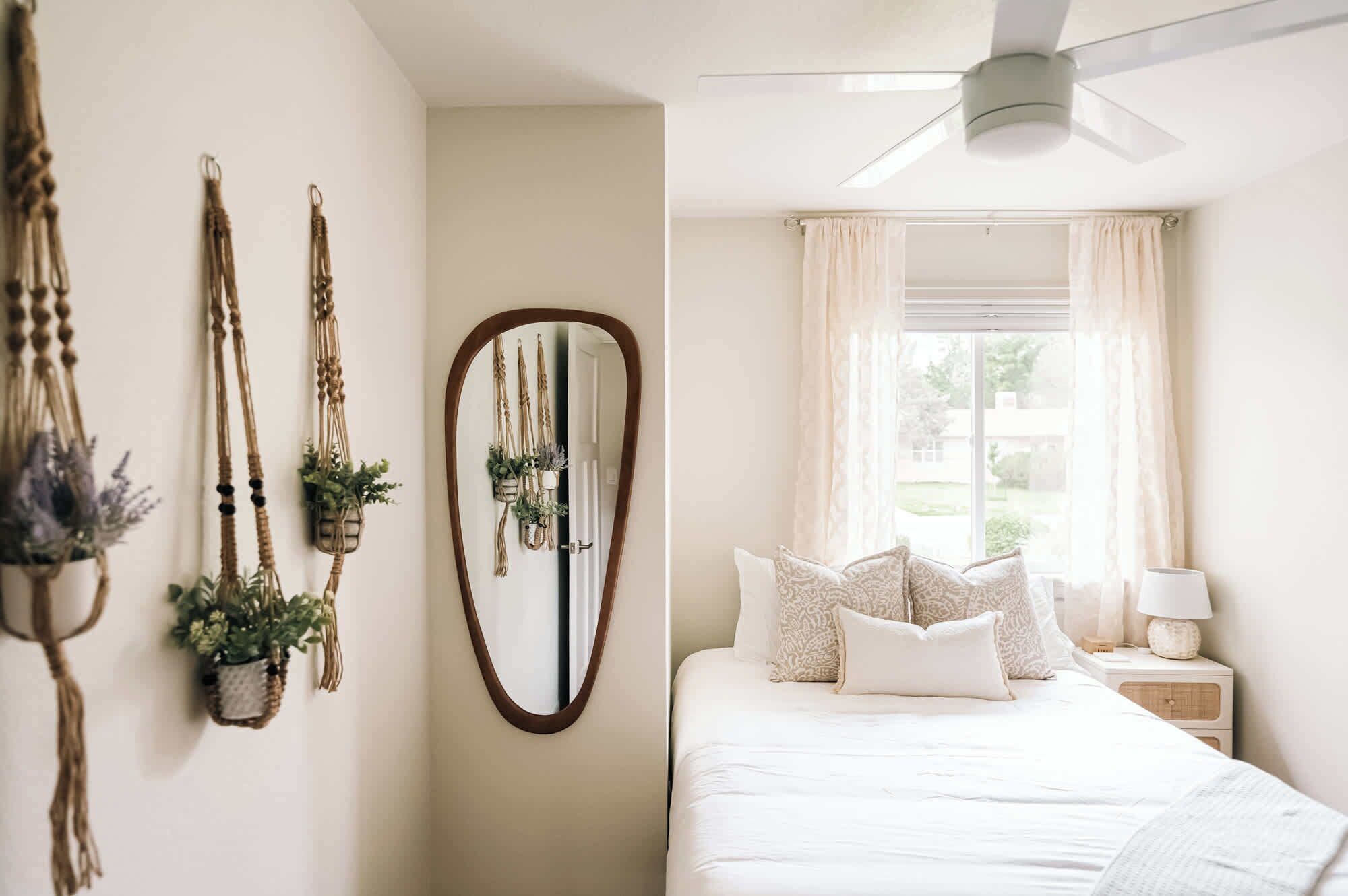 Ein Schlafzimmer mit Bett, Nachttisch, Deckenventilator, einem Fenster mit Vorhängen und wandmontierten Makramee-Dekorationen.