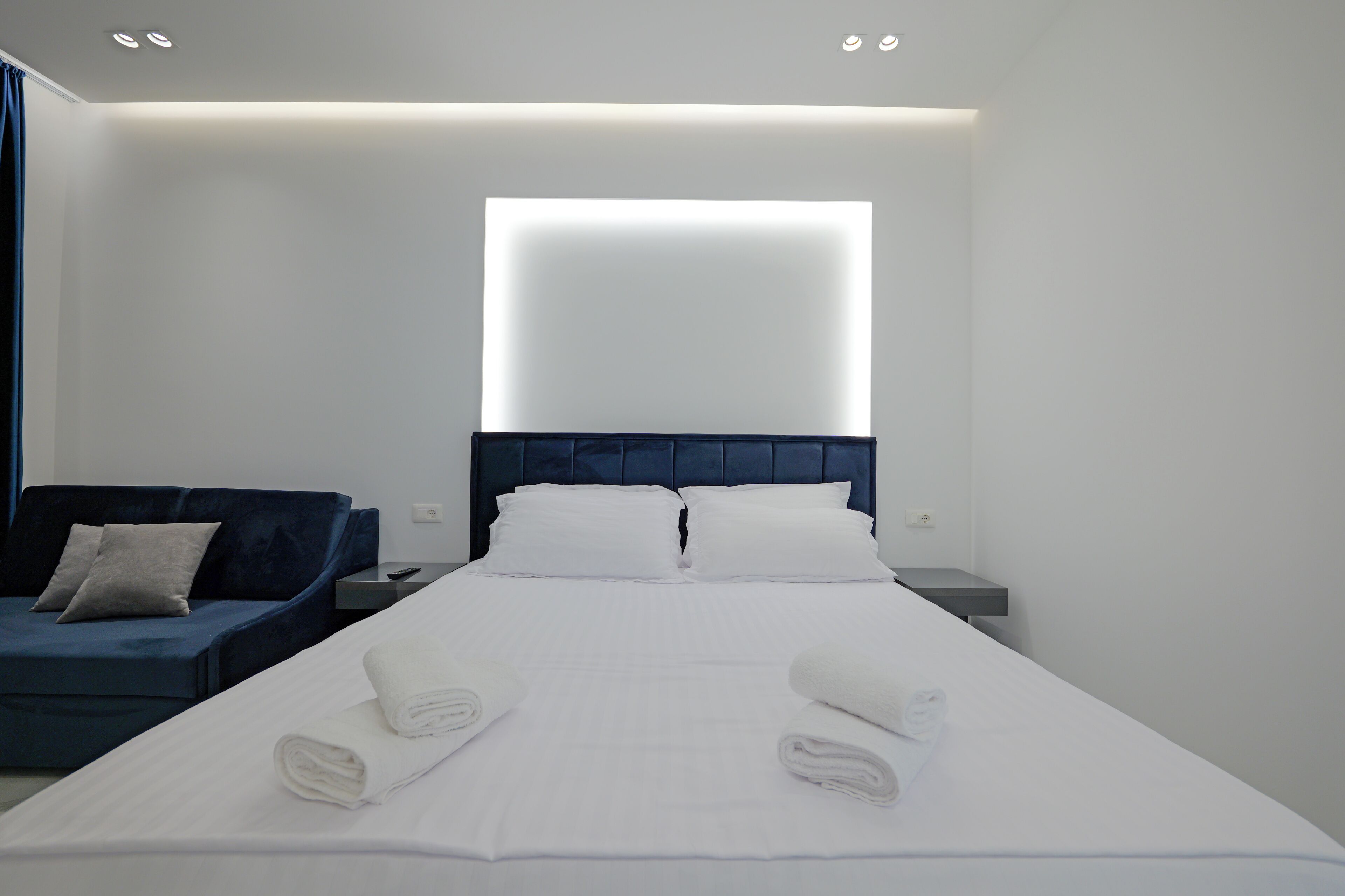 Una camera d'albergo moderna con un letto rifatto, un divano blu imbottito e un'ampia finestra con luce intensa.