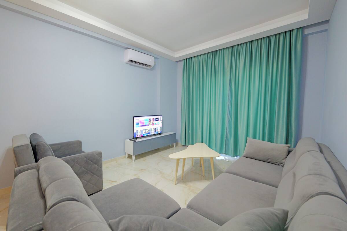 Una sala moderna con un sofá seccional gris, una mesita auxiliar blanca y un televisor de pantalla plana sobre un mueble gris.