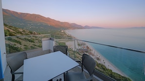Een balkon met een tafel en stoelen, uitkijkend op een kustlandschap met bergen en een strand.