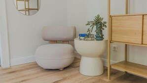 Une pièce avec un plancher en bois, un miroir rond, une chaise grise, un pouf blanc, une petite table blanche avec une plante et une étagère en bois.