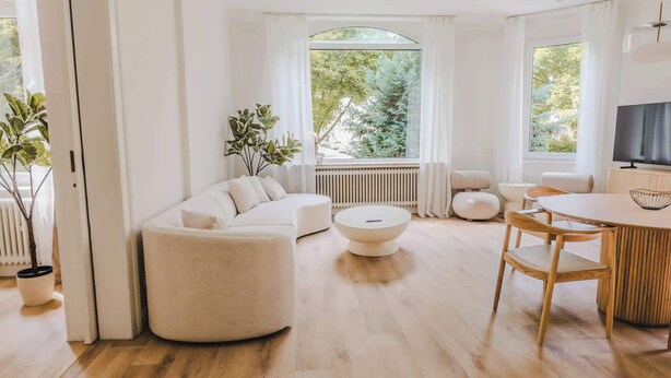 Un salon spacieux avec un canapé blanc, une table basse ronde et une table à manger en bois avec des chaises. Une grande fenêtre offre une vue sur la verdure environnante.
