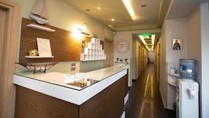 Zona de recepción de un hotel con mostrador, un reloj, estante en la pared con decoración de velero y dispensador de agua.