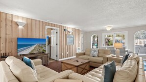 En rummelig stue med et stort fladskærms-tv, lysebeige sofaer, et træsofabord og havudsigt gennem vinduerne.