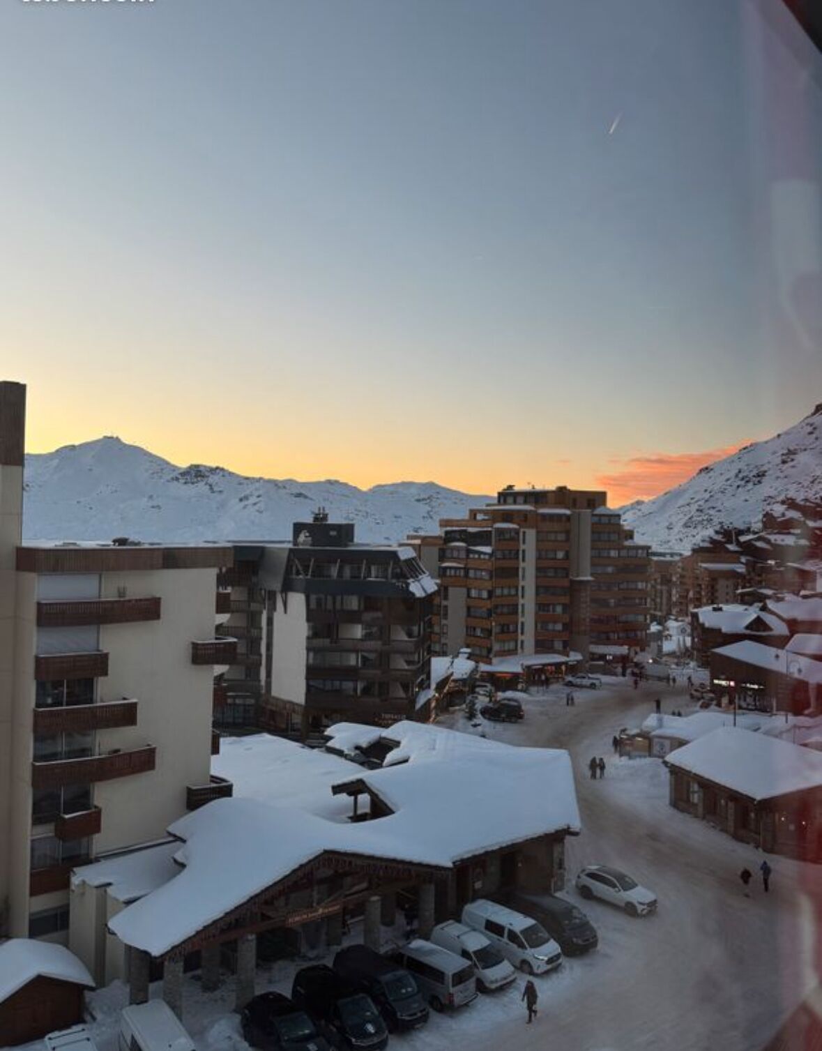En snötäckt stad med flera byggnader, inklusive ett hotell med balkonger och en parkeringsplats.
