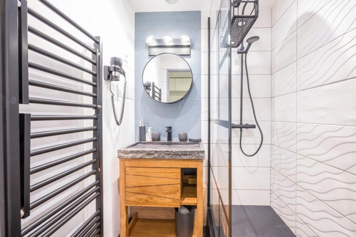 Une salle de bain avec un meuble-lavabo en bois, un miroir rond avec éclairage intégré, un espace douche et un mur carrelé.