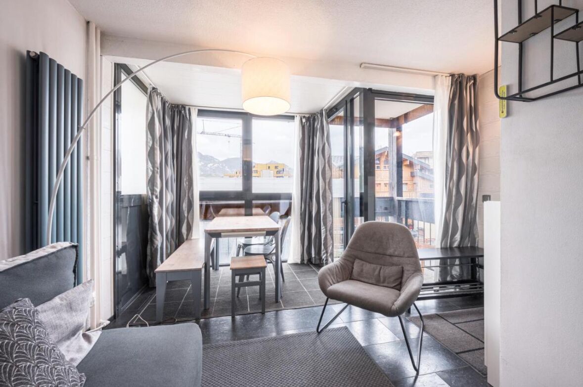 A modern living room with a sofa, a chair, a dining table, and a view through large windows.
