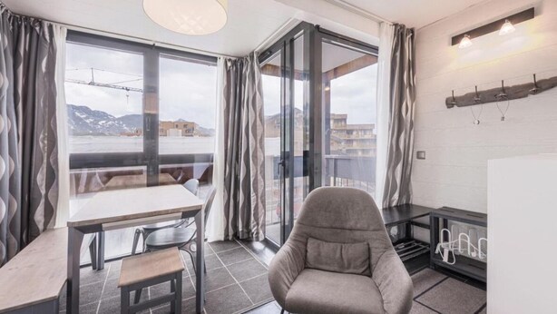 Une pièce moderne comprenant une table et des chaises, un canapé, une grande fenêtre avec des rideaux et un luminaire fixé au mur.