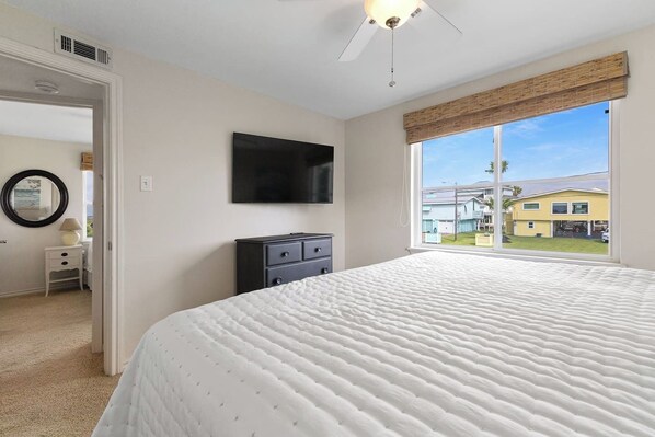 A bedroom with a large bed, a flat-screen TV, a dresser, and a window with a view of a yellow house.
