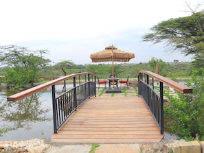 A wooden bridge leading to a table and chairs by a body of water, surrounded by greenery. - Esidai by Camp David (Nairobi)