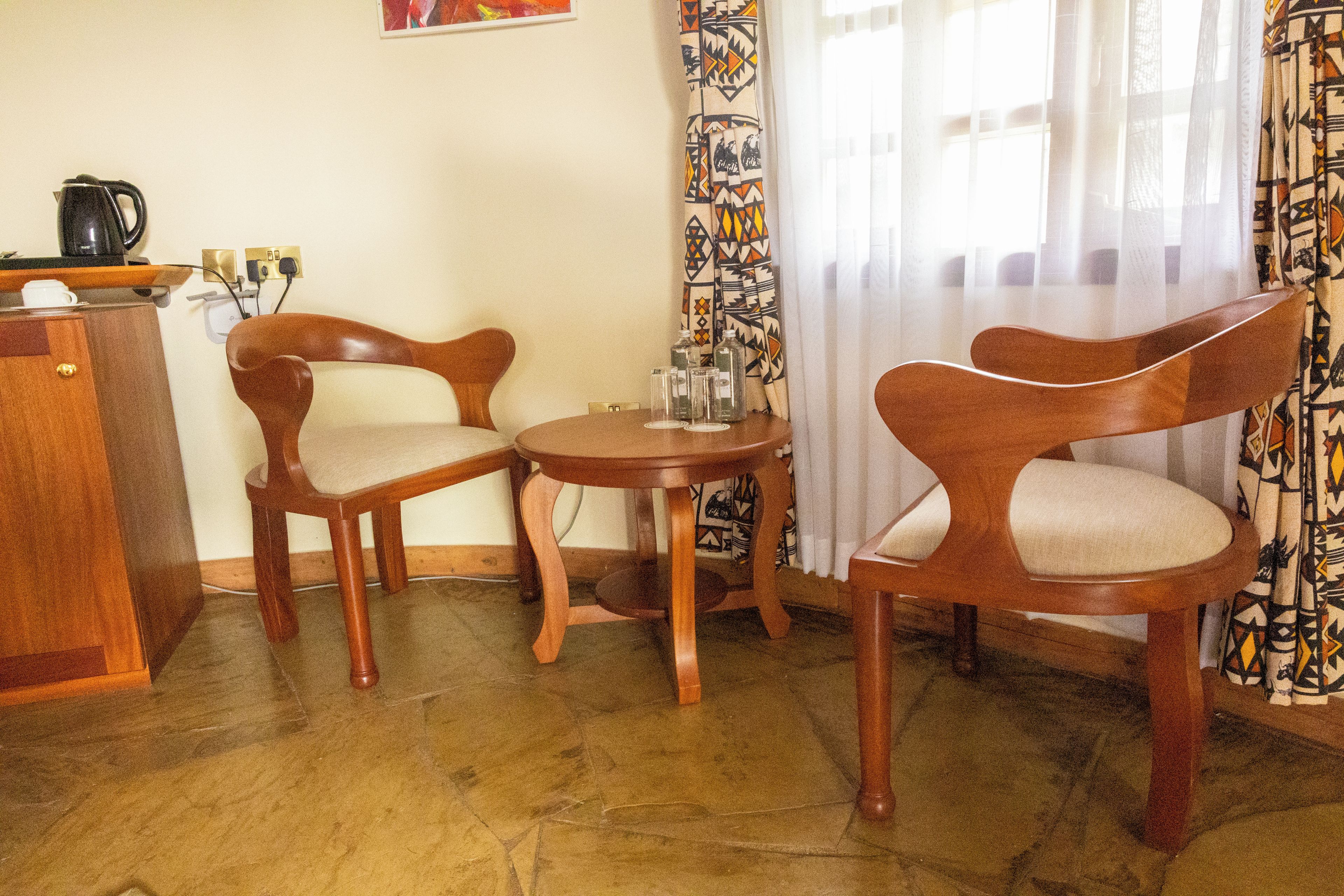 Una habitación con dos sillas de madera, una mesa redonda pequeña, una cafetera y cortinas estampadas.