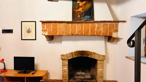 A room with a brick fireplace, a framed painting above it, a desk with a computer, and a bookshelf. - Casa Nerina (Castiglione d'Orcia)