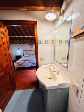 A narrow hallway leading to a bedroom with a bed, a bathroom with a sink and mirror, and a wooden ceiling. - Casa Nerina (Castiglione d'Orcia)