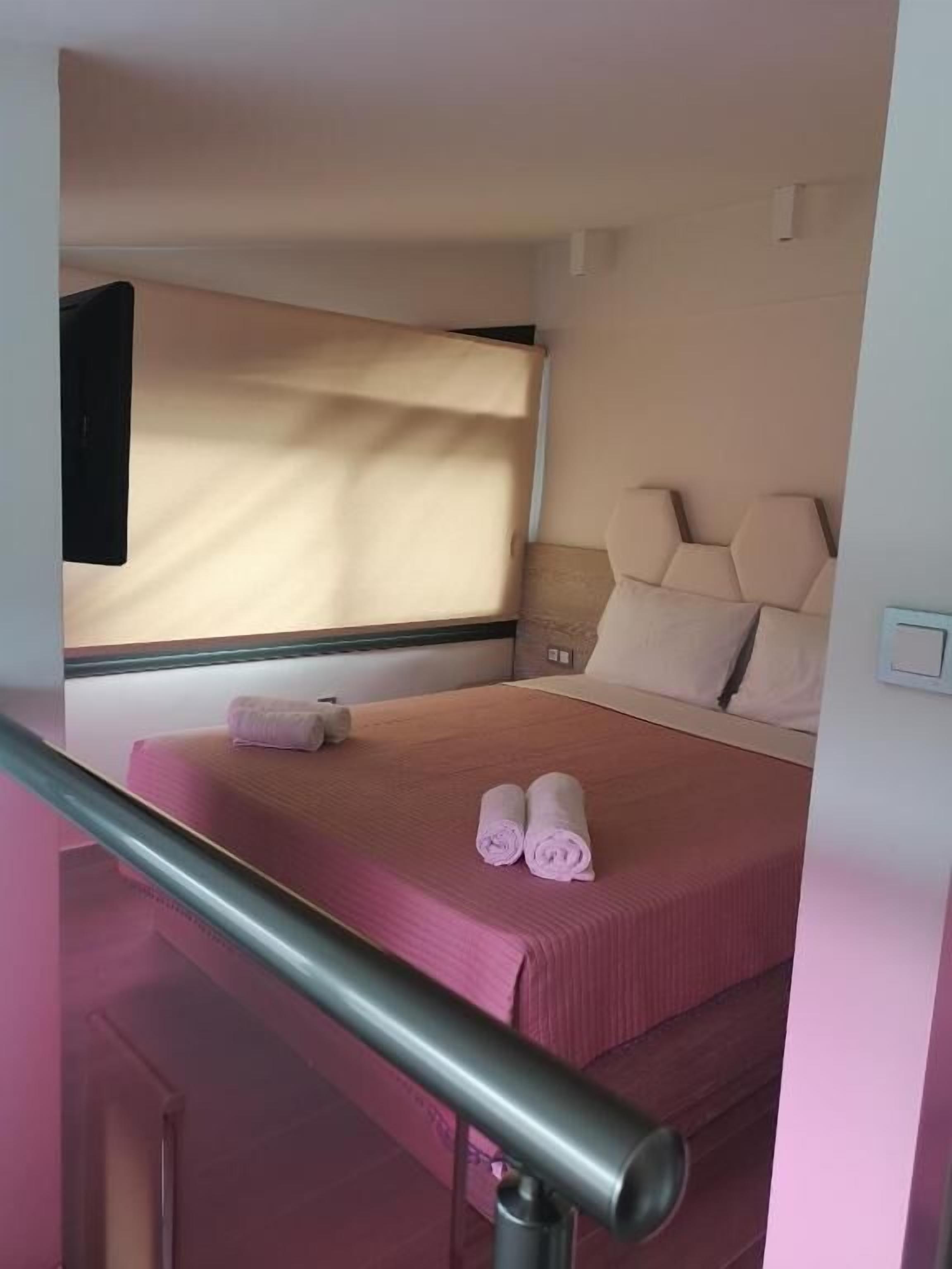 Ein kleines Hotelzimmer mit Bett, Fenster mit Vorhängen, Nachttisch und einem an der Wand befestigten Lichtschalter.