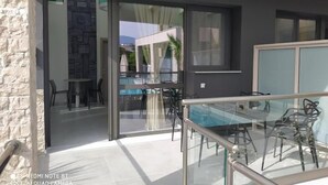 Een modern terras buiten met een glazen tafel en stoelen, een zwembad en een stenen muur.