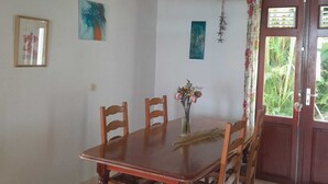Een eetgedeelte met een houten tafel en stoelen, een vaas met bloemen, en een deur met uitzicht op groen.