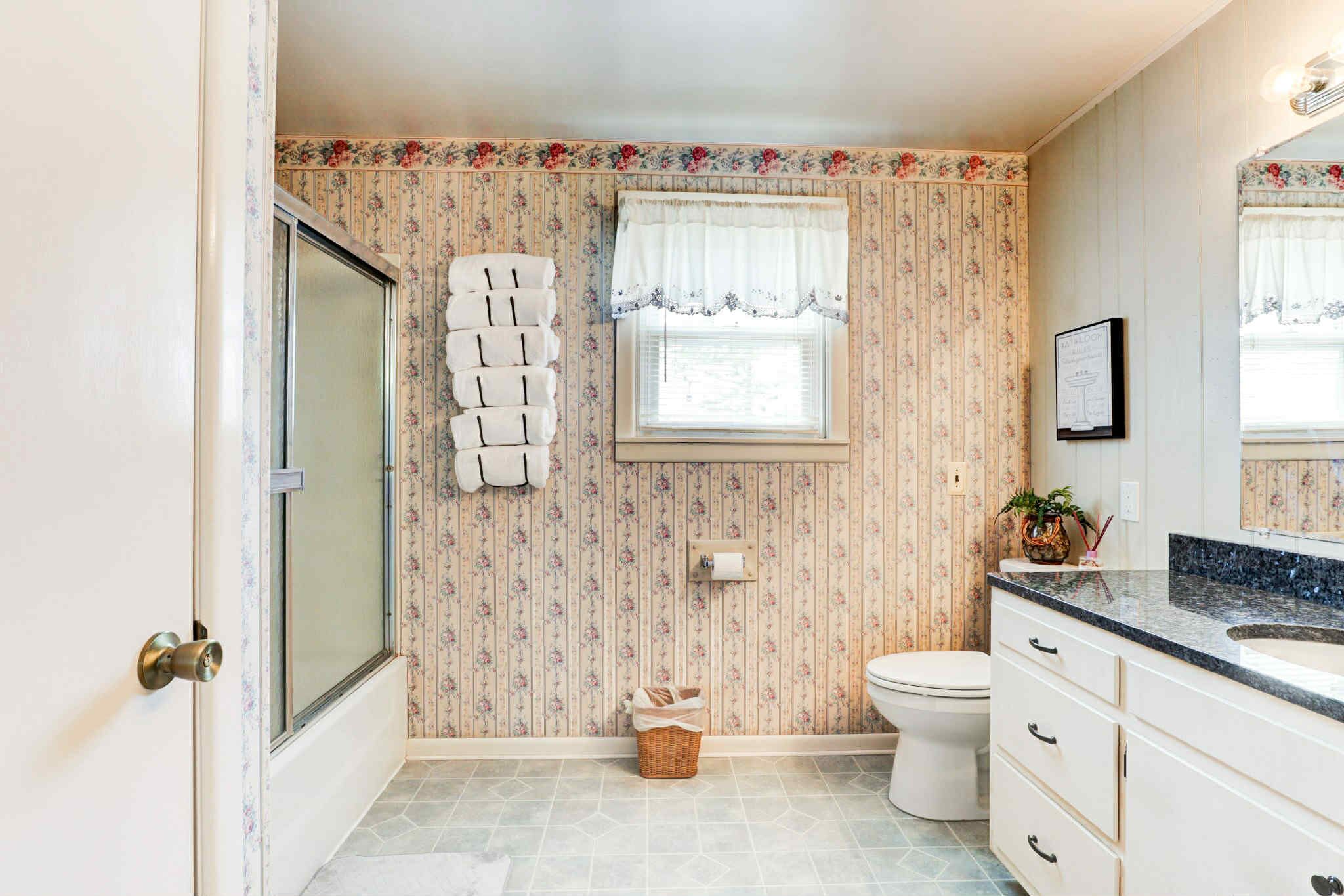 A bathroom with floral wallpaper, a toilet, a sink with a granite countertop, and a window with a white valance.