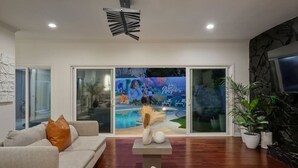 A modern living room with a sofa, a coffee table, and a TV. There is a large sliding glass door leading to an outdoor area with a pool and lounge chairs. - Blue Haven | Solar Pool Heat + Parking + Billiards (Long Beach)