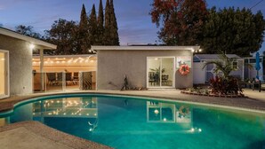 A swimming pool with a lifebuoy and a first aid station sign, surrounded by a building with a covered patio and outdoor seating. - Blue Haven | Solar Pool Heat + Parking + Billiards (Long Beach)