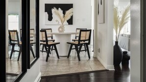 A dining area with a white table and four chairs, a vase with pampas grass, and a black and white artwork on the wall. - Blue Haven | Solar Pool Heat + Parking + Billiards (Long Beach)