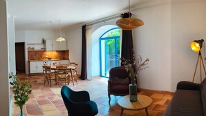 Amplia sala de estar con sofá, una mesa de comedor con sillas, cocina al fondo y un ventanal con cortinas.