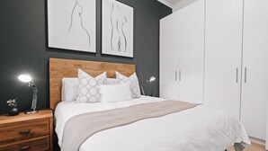 A modern bedroom with a wooden headboard, white bedding, and two wall-mounted artworks.