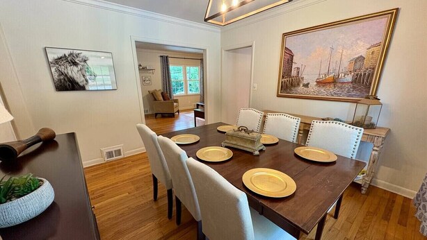 Uma sala de jantar com uma mesa de madeira posta para seis pessoas, rodeada por seis cadeiras estofadas. Há um quadro na parede, um abajur e uma pequena mesa lateral com uma planta e um objeto decorativo.