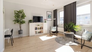 Un salon moderne avec un téléviseur à écran plat, une bibliothèque, une plante en pot et une fenêtre avec des rideaux.