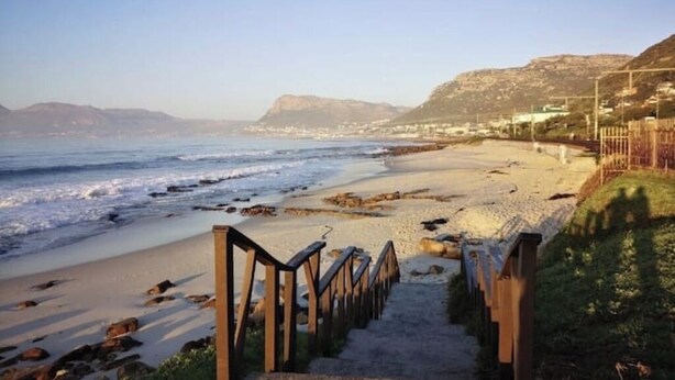 A beach with a wooden staircase leading down to the shore, surrounded by rocky terrain and hills in the background. - Kalk Bay Secure Seaside Apartment (Cape Town)