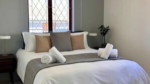 A neatly made bed with white and gray linens, a gray throw blanket, and two rolled towels. There is a small bedside table with a lamp and a potted plant. - Kalk Bay Secure Seaside Apartment (Cape Town)