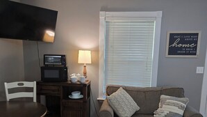 Una sala de estar con piso de madera, un sofá gris con dos cojines, una mesita con una lámpara, un microondas y un televisor sobre un mueble.