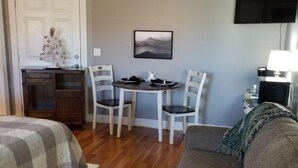 Una habitación acogedora con una cama, un sofá gris, una mesa de comedor con sillas, un escritorio pequeño, un televisor y una obra de arte colgada en la pared.
