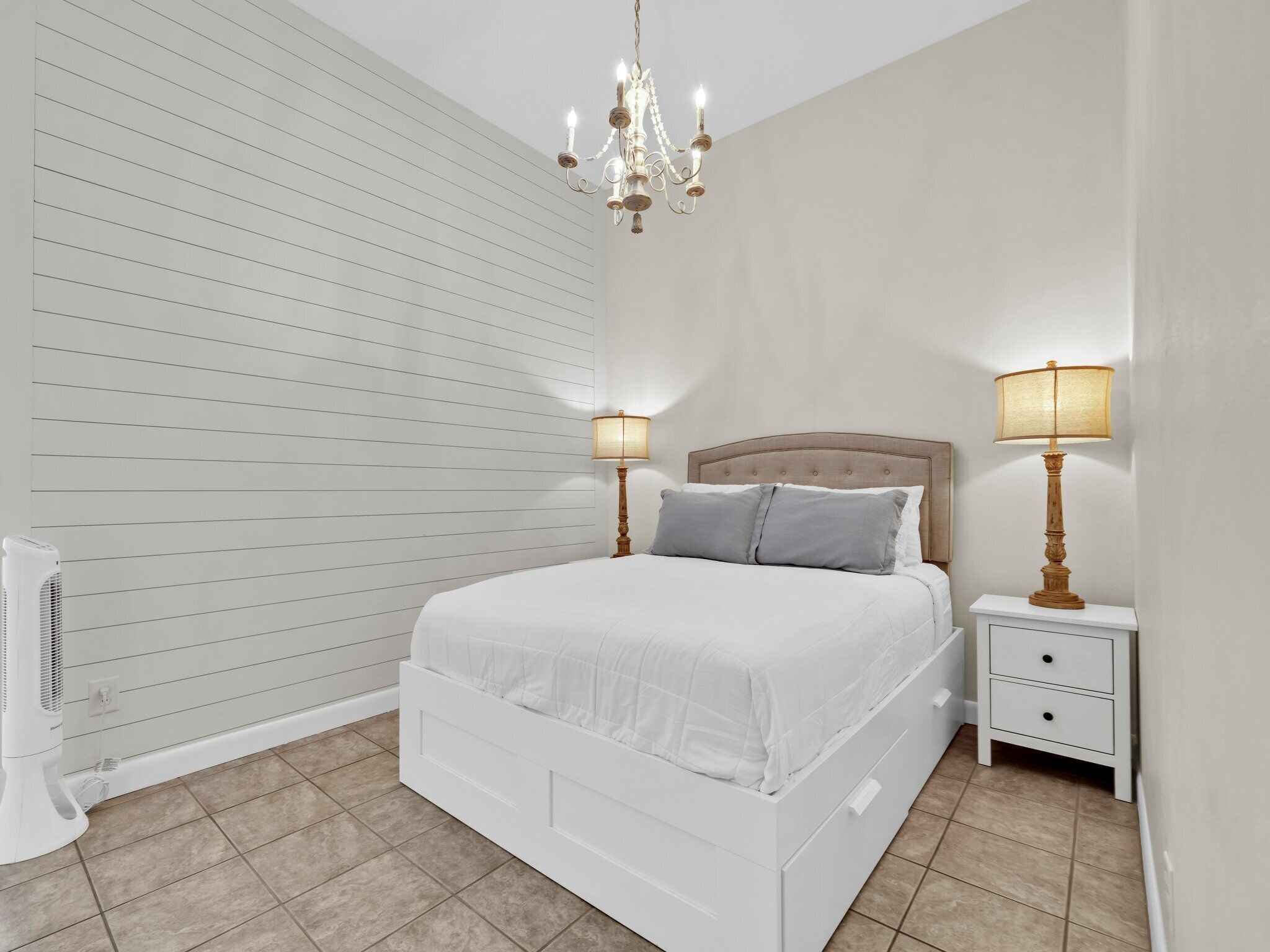 Une chambre à coucher avec un lit blanc, deux tables de chevet avec des lampes, un ventilateur de plafond et un lustre.