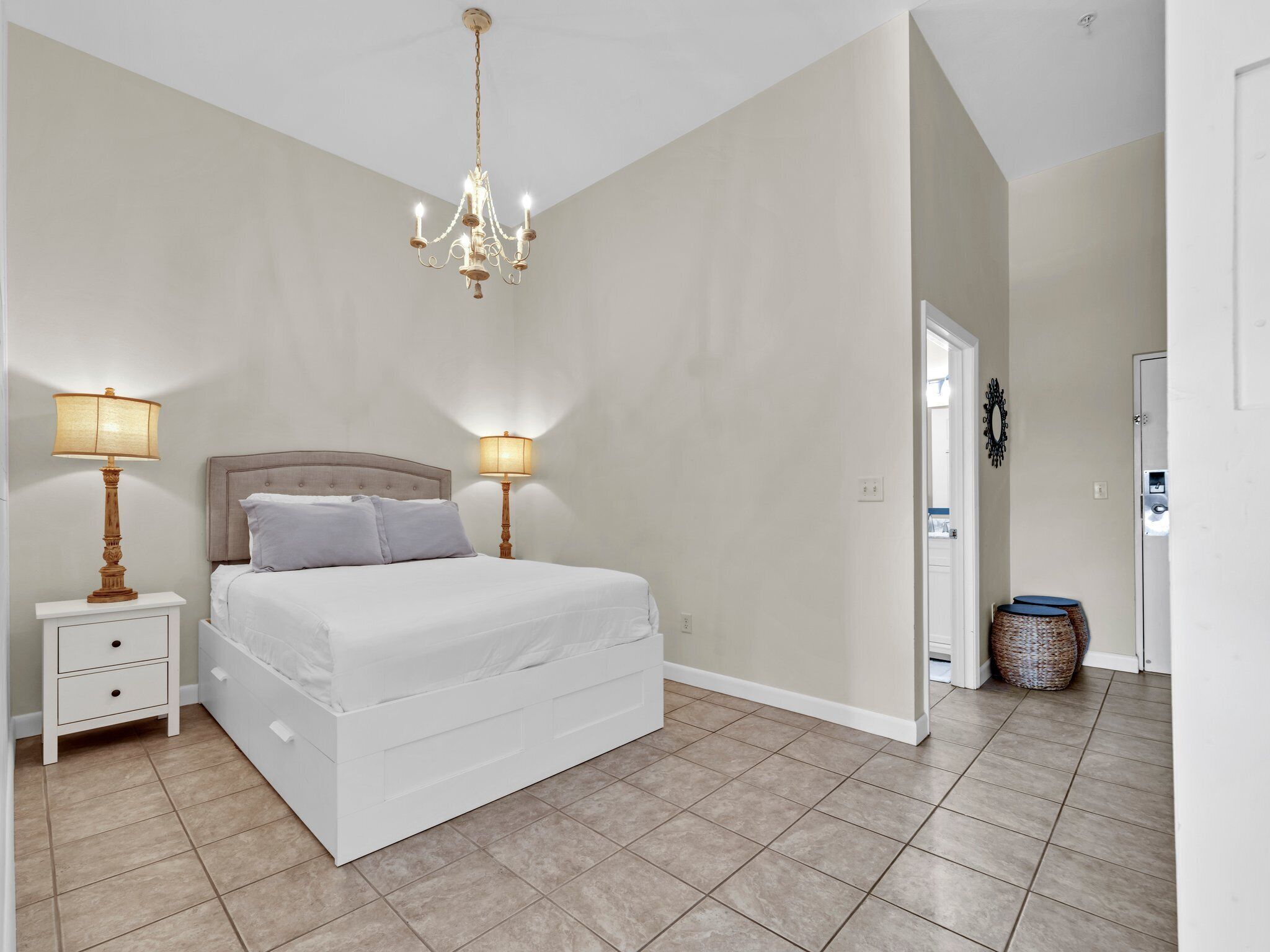 A bedroom with a large bed, two bedside tables with lamps, a chandelier, and a door leading to another room.