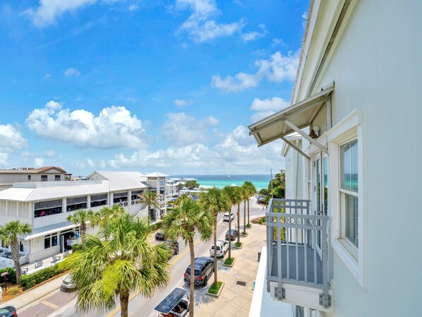 A coastal hotel with a balcony overlooking a street lined with palm trees and parked cars, leading to a beach.