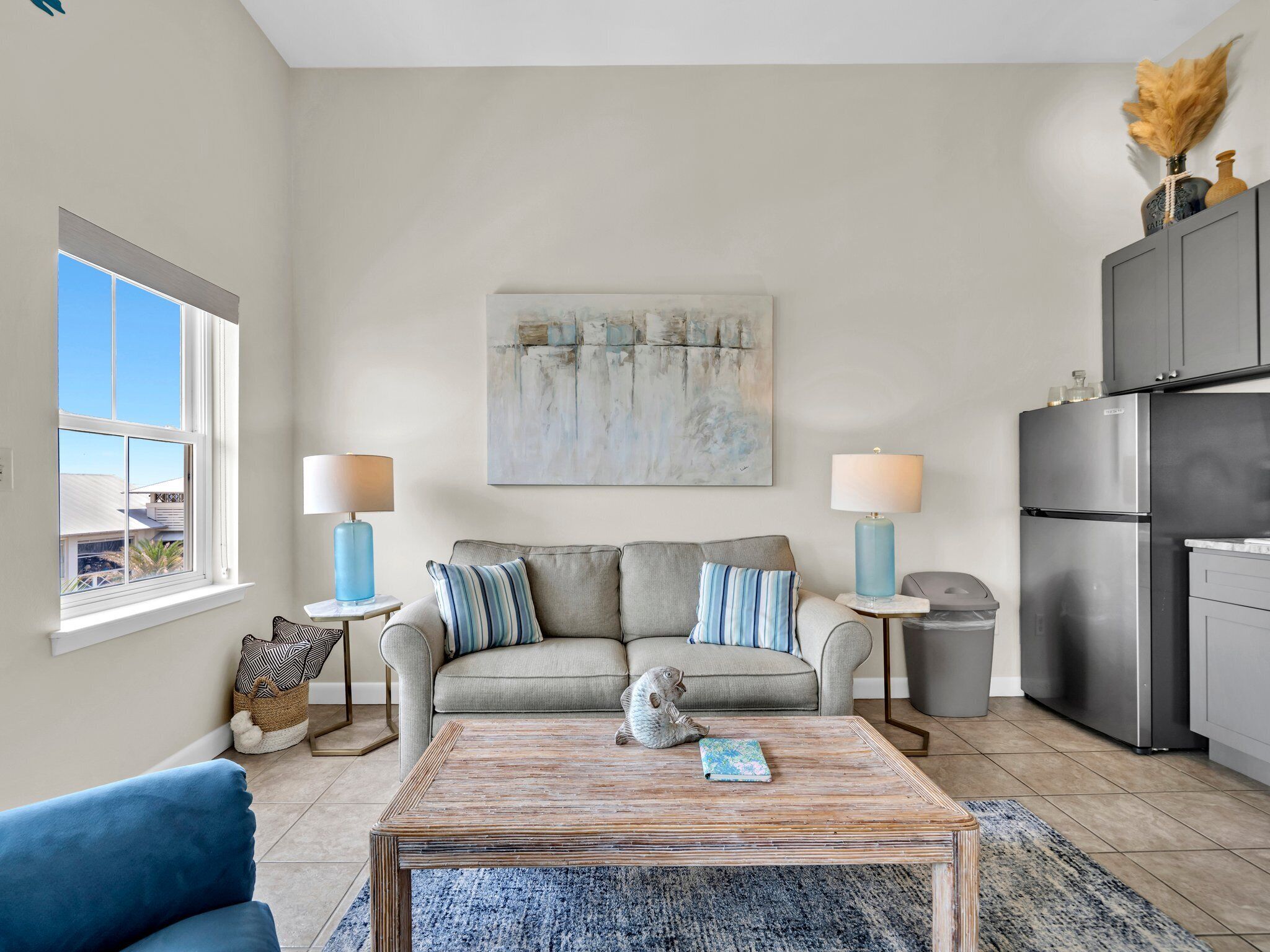 A modern living room with a sofa, two side tables with lamps, a coffee table, and a kitchen area with a refrigerator.