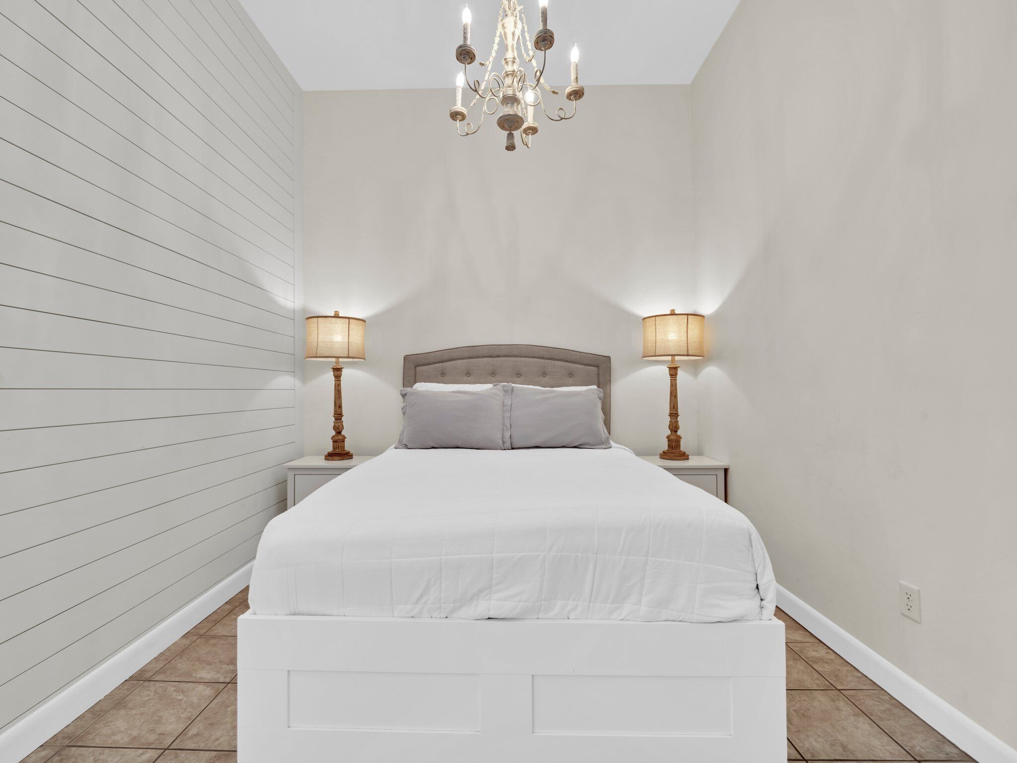 A bedroom with a large bed, two bedside tables with lamps, a chandelier, and a white wall with horizontal lines.