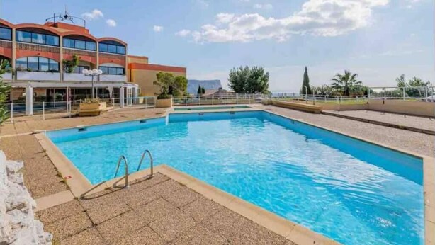 Une piscine à l’eau d’un bleu limpide, entourée d’une zone pavée et d’un bâtiment doté d’un toit rouge et de nombreuses fenêtres.