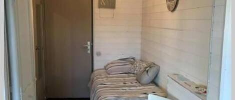 Una habitación pequeña y funcional con cama, silla, escritorio y una lámpara colgante.