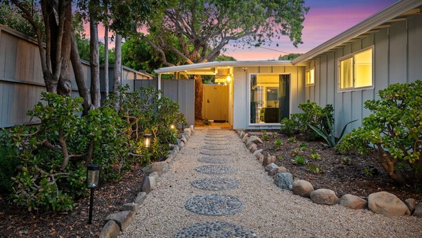 A pathway leading to a house with a covered entryway, surrounded by trees and a gravel path.
