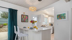 A modern kitchen with a central island, white bar stools, and a dining area with a view of the outdoors.