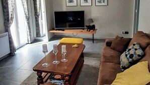Een moderne woonkamer met een bruine bank, een geel poefje en een houten salontafel met drie wijnglazen. Een flatscreen televisie is aan de muur bevestigd, en erboven hangen drie ingelijste foto’s.