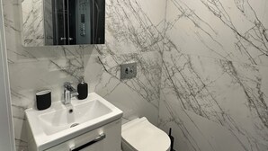 Un baño moderno con pared de mármol blanco, un lavamanos blanco, un inodoro y un espejo sobre el lavamanos.