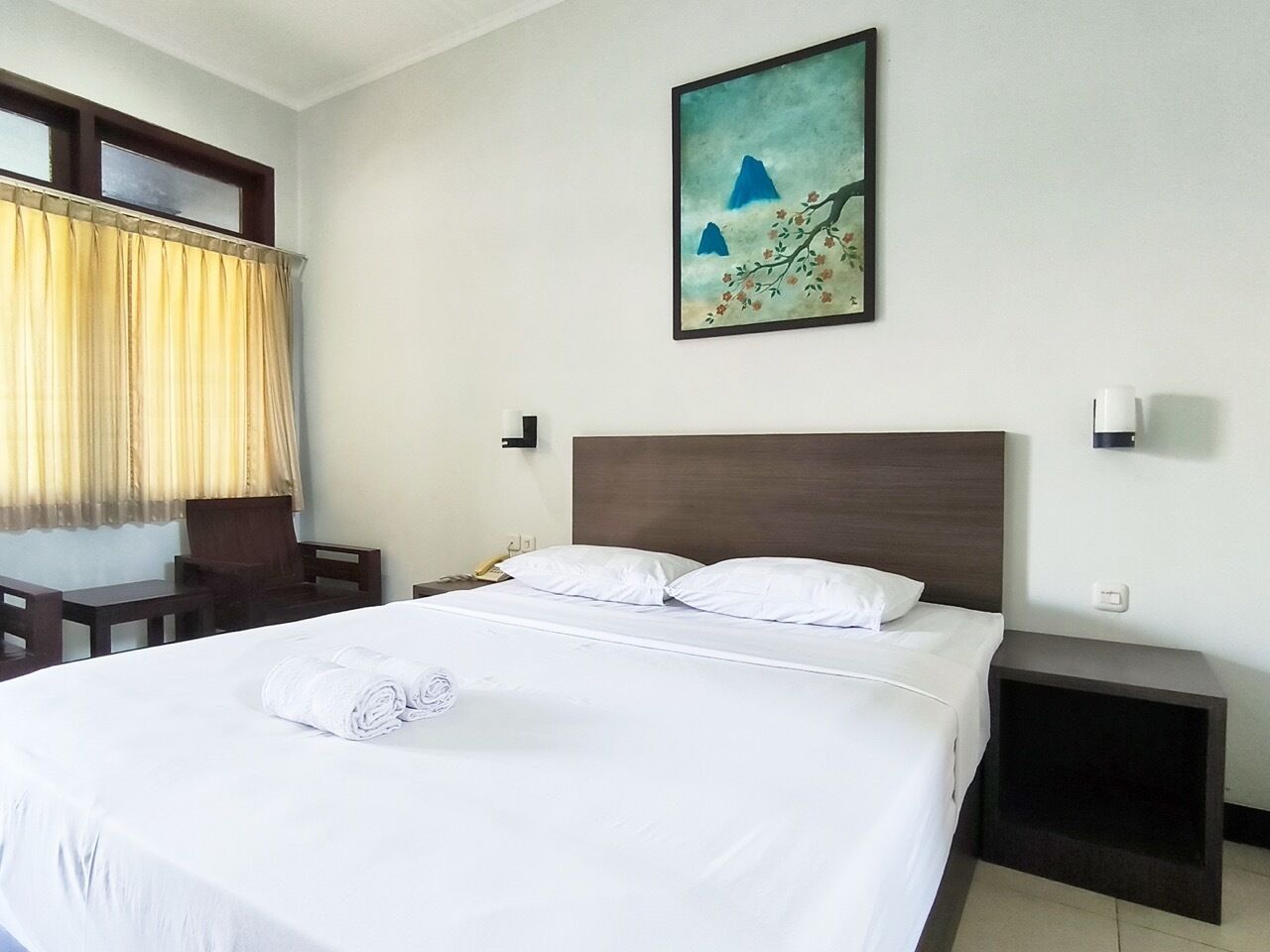 A hotel room with a large bed, a wooden headboard, a painting on the wall, a bedside table, and a chair.