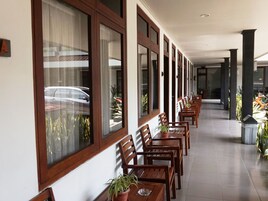 A hotel corridor with wooden chairs and tables, large windows, and potted plants.