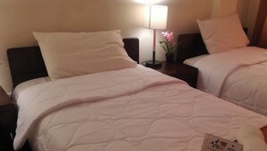 A hotel room with two beds, each with a white quilted bedspread and matching pillows. There is a nightstand with a lamp and a small potted plant. A welcome card is placed on the bed. - hola wellness  (Cusco)